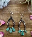 Pair of beaded earrings with turquoise teardrop charms on a wooden surface with floral decorations.