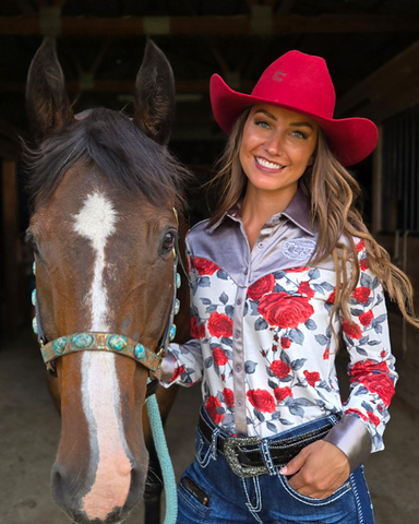 Woman in a red cowboy hat and floral shirt standing next to a horse in a barn.