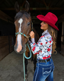 Woman in a red cowboy hat and floral shirt standing next to a horse in a stable.