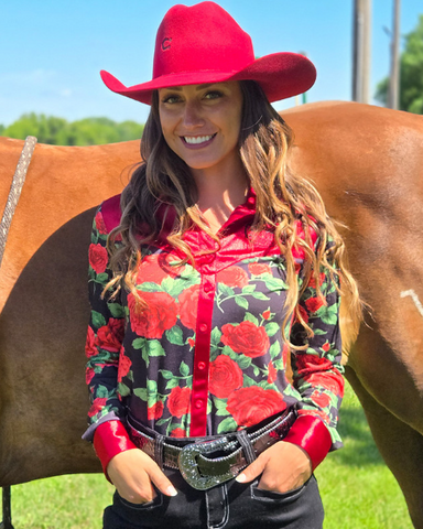 Woman in a red cowboy hat and floral shirt standing next to a horse outdoors.