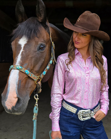 Woman in pink shirt and cowboy hat standing next to a horse in a stable.