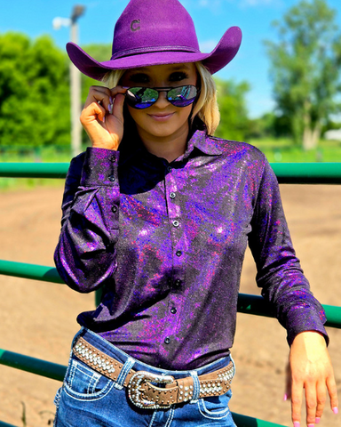 Person wearing a purple cowboy hat and shirt with a glittery texture, standing in an outdoor setting.