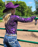 Person wearing a purple iridescent shirt and cowboy hat, standing in a fenced area with trees in the background.