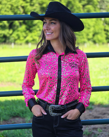 Woman wearing a pink and leopard patterned shirt with a black cowboy hat in an outdoor setting.