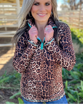 A woman wearing a leopard print zip-up fleece jacket with turquoise zipper pulls, standing outdoors in front of a wooden fence and greenery.