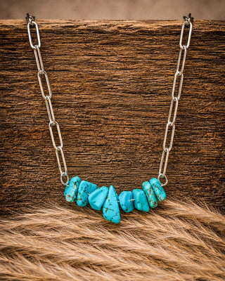 A necklace with a curved row of polished turquoise-colored stone chips on a silver paperclip chain, displayed on a rustic wooden surface with tan fur in the foreground.