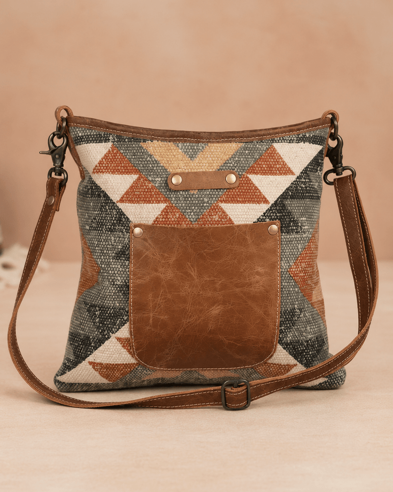 A geometric patterned crossbody handbag with a brown faux-leather front pocket and matching brown strap, set against a warm beige background.