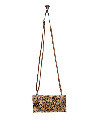 A brown leather crossbody bag with an intricate hand-tooled floral pattern, featuring a long adjustable brown leather strap and a top hanging hook, shown against a plain white background.