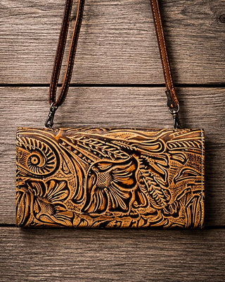 A brown tooled leather clutch bag with an intricate floral and scroll pattern, featuring a matching leather shoulder strap, displayed flat on a rustic wooden plank background.