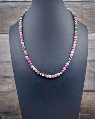 A beaded necklace displayed on a black jewelry bust against a rustic wood plank background. The necklace features round beads in pink, purple, orange, white, and metallic silver.