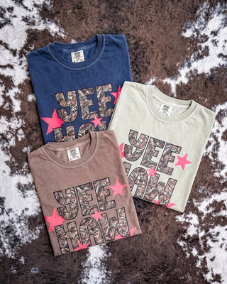 Three folded graphic t-shirts in navy blue, espresso brown, and light khaki, laid out on a brown and white cowhide rug. Each shirt features a camouflage print graphic reading 'YEEHAW' and is decorated with red star accents.