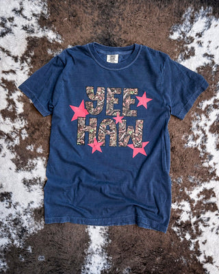 A navy short-sleeve t-shirt laid flat on a brown and white cowhide rug. The shirt features a graphic print reading 'YEE HAW' in a camouflage pattern, surrounded by red stars.