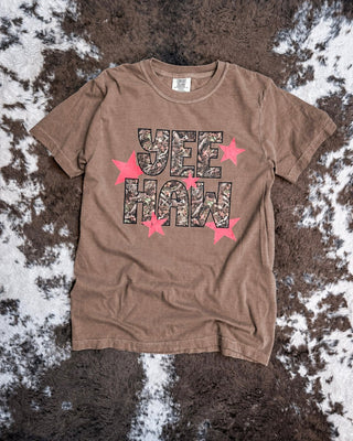 A brown short-sleeve t-shirt laid flat on a brown and white cowhide rug. The shirt features a graphic print reading 'YEE HAW' in a camouflage pattern, surrounded by red stars.