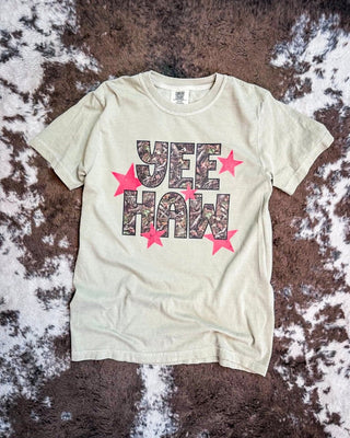 A khaki short-sleeve t-shirt laid flat on a brown and white cowhide rug. The shirt features a graphic print reading 'YEE HAW' in a camouflage pattern, surrounded by red stars.