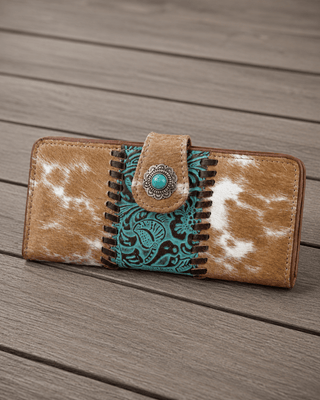 A western-style long wallet with a tan and white cowhide pattern, a turquoise and black paisley embroidered center panel, and a decorative metal snap button, shown on a gray wooden plank surface.