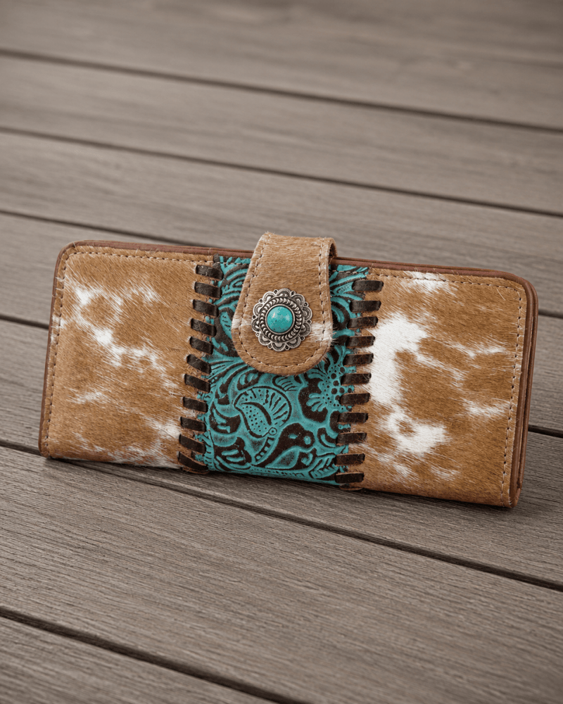 A western-style long wallet with a tan and white cowhide pattern, a turquoise and black paisley embroidered center panel, and a decorative metal snap button, shown on a gray wooden plank surface.