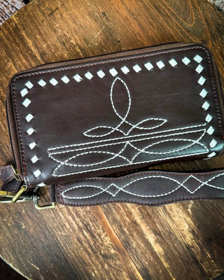 A dark brown leather zip-around wallet with white decorative stitching, resting on a rustic wooden table. The wallet features a geometric diamond border and a central abstract stitched design, with a matching wrist strap attached.