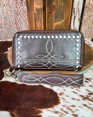 A brown leather zip-around wallet with white decorative stitching, silver studs, and a matching wristlet strap, displayed on a wooden stand over a cowhide rug.