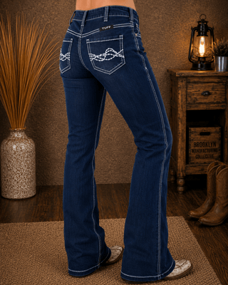 A person shown from the waist down wearing dark blue western flare jeans with white contrast stitching and decorative stitching on the back pockets, standing in a rustic room with a wooden side table, boots, and a lamp.