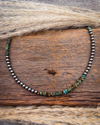 A beaded necklace featuring metallic silver beads and turquoise-colored stone chips, displayed on a rustic wooden surface with dried grass.