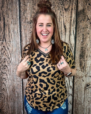 A woman smiling and posing in front of a rustic wood plank wall, wearing a short-sleeve leopard print top in tan and black with blue jeans.