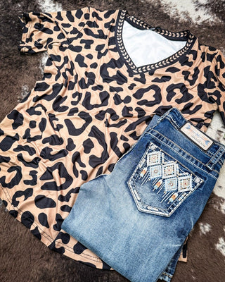 A beige and black leopard print V-neck t-shirt with decorative trim, styled flat-lay with folded blue jeans featuring a geometric embroidered pocket on a brown and white cowhide rug.