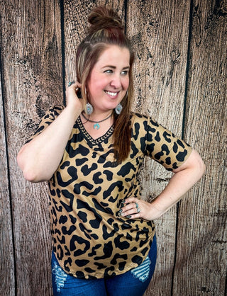 A woman wearing a leopard print V-neck short-sleeve top and blue jeans, posing in front of a rustic wood plank background.