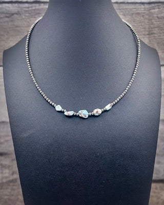 A silver beaded necklace with five iridescent, irregularly shaped blue-green stones, displayed on a black jewelry bust against a rustic gray wood background.