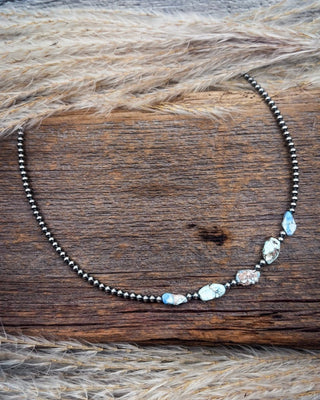 A beaded necklace with dark metallic beads and irregular light blue and white stones, displayed on a rustic wooden surface with dried grass.
