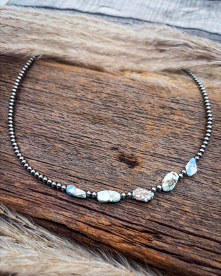 A beaded necklace with small dark metallic beads and irregular natural stone accents in light blue, white, and brown, displayed on a rustic wooden surface with tan fur.