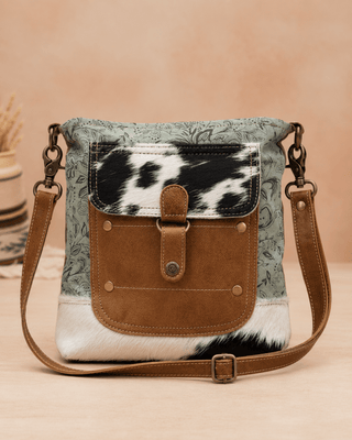 A western-style crossbody bag with a sage green floral patterned body, a black and white cowhide flap, a brown suede front pocket, and a matching brown shoulder strap, set against a warm beige background.
