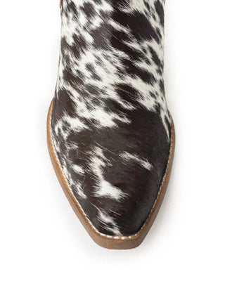 A close-up top-down view of a pointed-toe boot featuring a black and white cowhide pattern, with a light brown sole, set against a plain white background.
