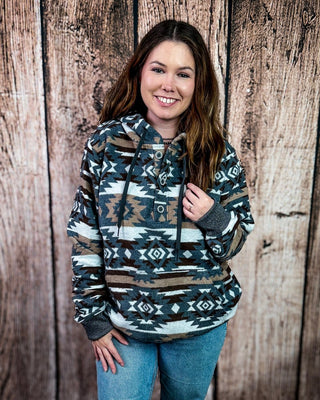 A woman smiling while wearing a long-sleeve hooded pullover with a Southwestern geometric pattern in brown, beige, gray, and white, paired with blue jeans, standing against a rustic wooden plank wall.