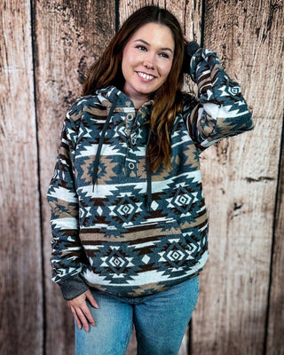 A woman wearing a pullover hoodie with a Southwestern geometric pattern in teal, brown, beige, and white, paired with blue jeans, standing against a rustic wooden plank wall.