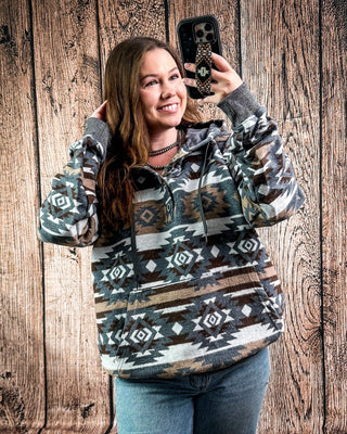 A woman takes a mirror selfie wearing a long-sleeve pullover hoodie with a Southwestern geometric pattern in gray, brown, beige, and black. She is standing in front of a rustic wooden plank wall.