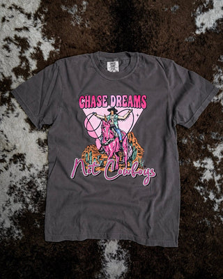 A dark gray graphic t-shirt laid flat on a brown and white cowhide rug. The shirt features a colorful graphic of a cowboy riding a horse with a heart outline, and the text 'CHASE DREAMS Not Cowboys' in pink and orange.