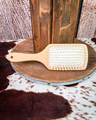 A wooden paddle hairbrush with white bristles rests on a round wooden platform over a cowhide rug, with a rustic wooden backdrop behind it.