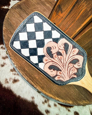 A hand-tooled leather hair brush with a black and white checkerboard design and a carved floral motif, resting on a rustic wooden surface.
