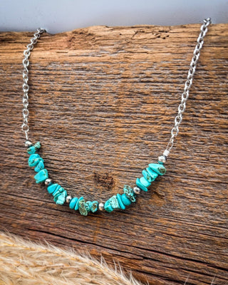 A silver chain necklace featuring a central row of irregular turquoise-colored stone beads and small metallic spacer beads, displayed on a rustic wooden surface.