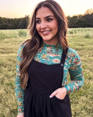 A woman standing in a grassy field at sunset, wearing black overall-style pants over a long-sleeve patterned top with a teal and tan botanical print.