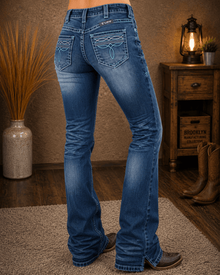 A person wearing blue western flare jeans with decorative embroidered details on the back pockets, shown from the waist down in a rustic indoor setting with a wooden cabinet, lamp, and boots.