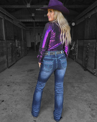 A woman stands in a barn aisle wearing a purple cowboy hat, a sparkly purple long-sleeve top, and blue jeans, shown from the back.