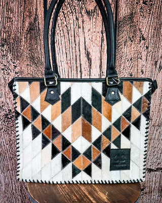 A structured tote bag with a geometric patchwork pattern of black, white, tan, and brown cowhide triangles, featuring black leather handles and trim, displayed on a wooden stool against a rustic wood plank background.