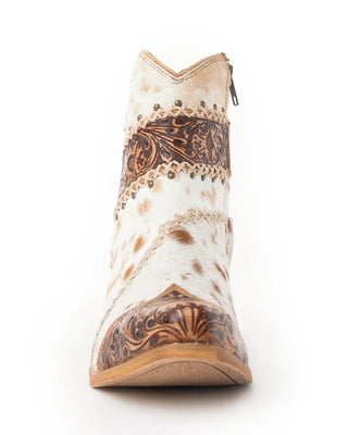 A single western-style ankle boot shown from the front on a white background. The boot features a white and tan cowhide pattern with dark brown tooled leather detailing around the collar, vamp, and quarter panels, and a small silver zipper at the top.