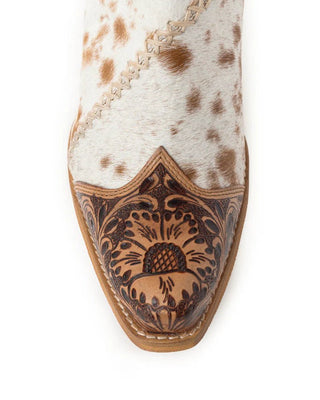 A close-up of a western boot's pointed toe showing a white and brown speckled hair-on-hide upper, contrast stitching, and a dark brown tooled leather vamp with floral scrollwork.