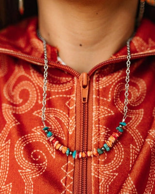 A close-up of a person wearing a silver chain necklace with a row of orange and turquoise beads, styled over a rust-orange patterned zip-up jacket.