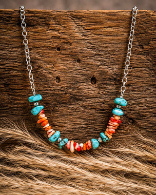 A bohemian-style necklace with turquoise and orange coral beads on a silver chain, displayed on a rustic wooden surface with dried grass.
