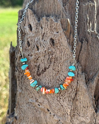 A beaded necklace with turquoise, orange, and white shell-like beads on a silver chain, displayed on a weathered tree stump outdoors.
