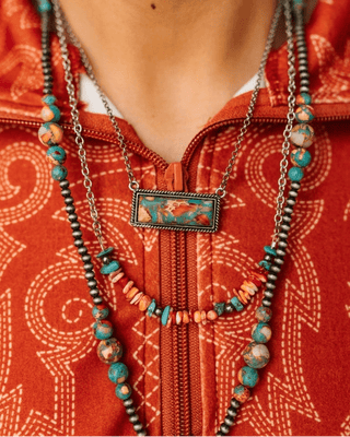 A close-up of a person wearing a layered bohemian necklace set over a rust-orange patterned top. The necklace features a central rectangular turquoise and gold pendant, accented by turquoise and orange beads, silver chains, and dark metallic spacer beads.