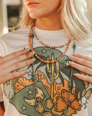 Person wearing a white t-shirt with a colorful graphic design, holding a beaded necklace.